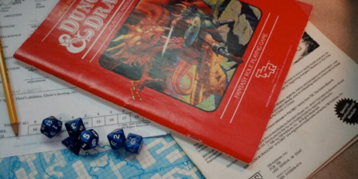 A vintage Dungeons & Dragons red box set, blue dice, character sheet, and pencil on a tabletop, with a map partially visible underneath.